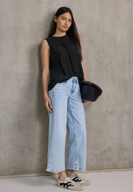 Frau mit schwarzer Bluse, heller Jeans und Sneakern steht vor Betonwand.